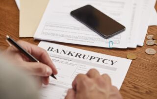 partial view of businessman filling in bankruptcy form at wooden table with smartphone