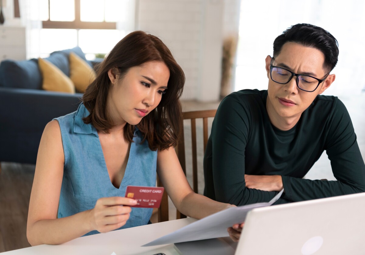 serious Asian wife checking analyzing statement utilities bills sitting together at home.